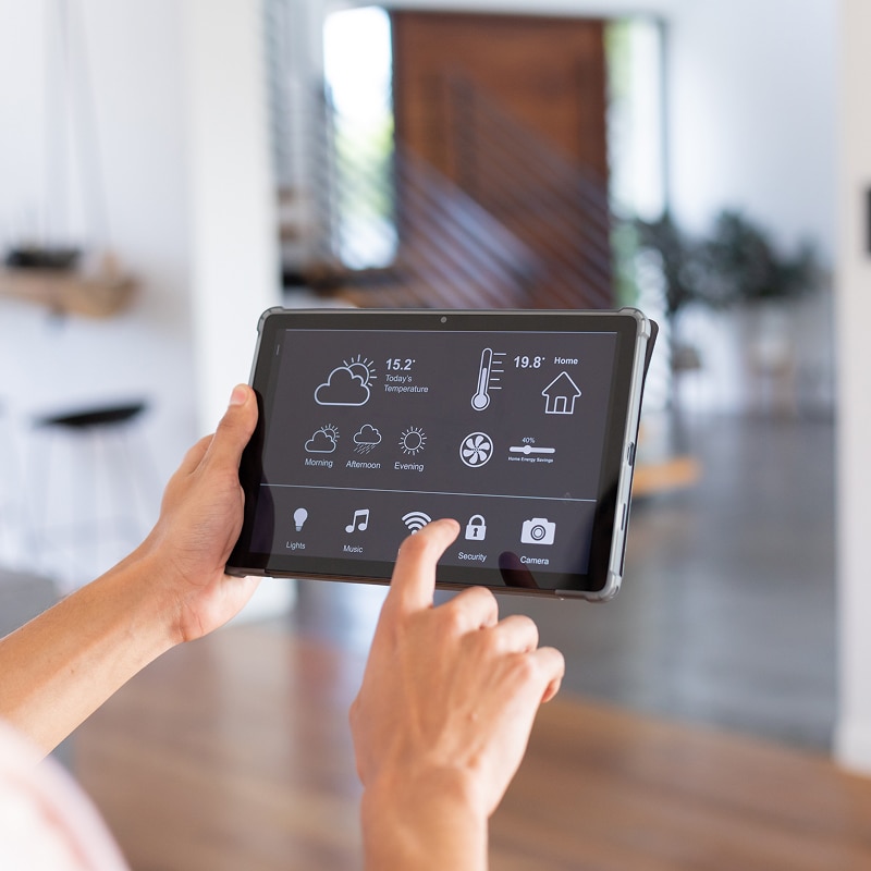 Main contrôle une tablette affichant une interface domotique : température (15.2°, 19.8°), météo, énergie (40% économies), lumières, musique, caméra. Un doigt touche "Security".