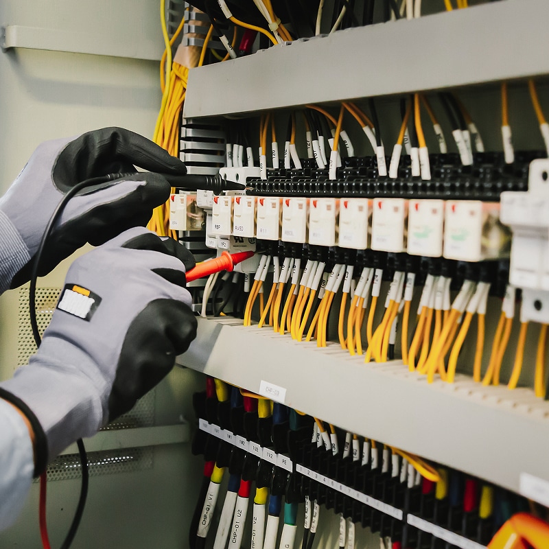 Un électricien en gants de sécurité teste des borniers d'un panneau électrique avec un multimètre, vérifiant le câblage jaune et étiqueté. Mentions: RBP, RA50, CHP-03.