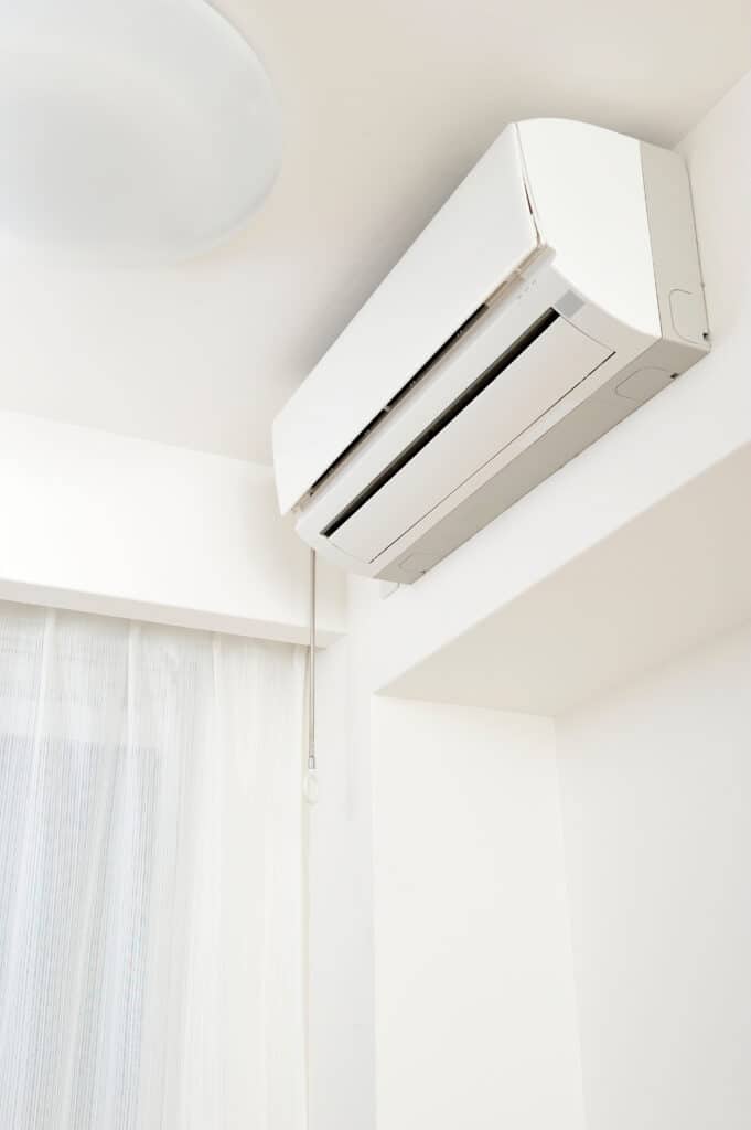 Climatiseur blanc mural : confort moderne dans la pièce Climatiseur mural blanc installé en hauteur dans une pièce lumineuse avec plafond et murs clairs. Fenêtre avec rideaux transparents rayés.
