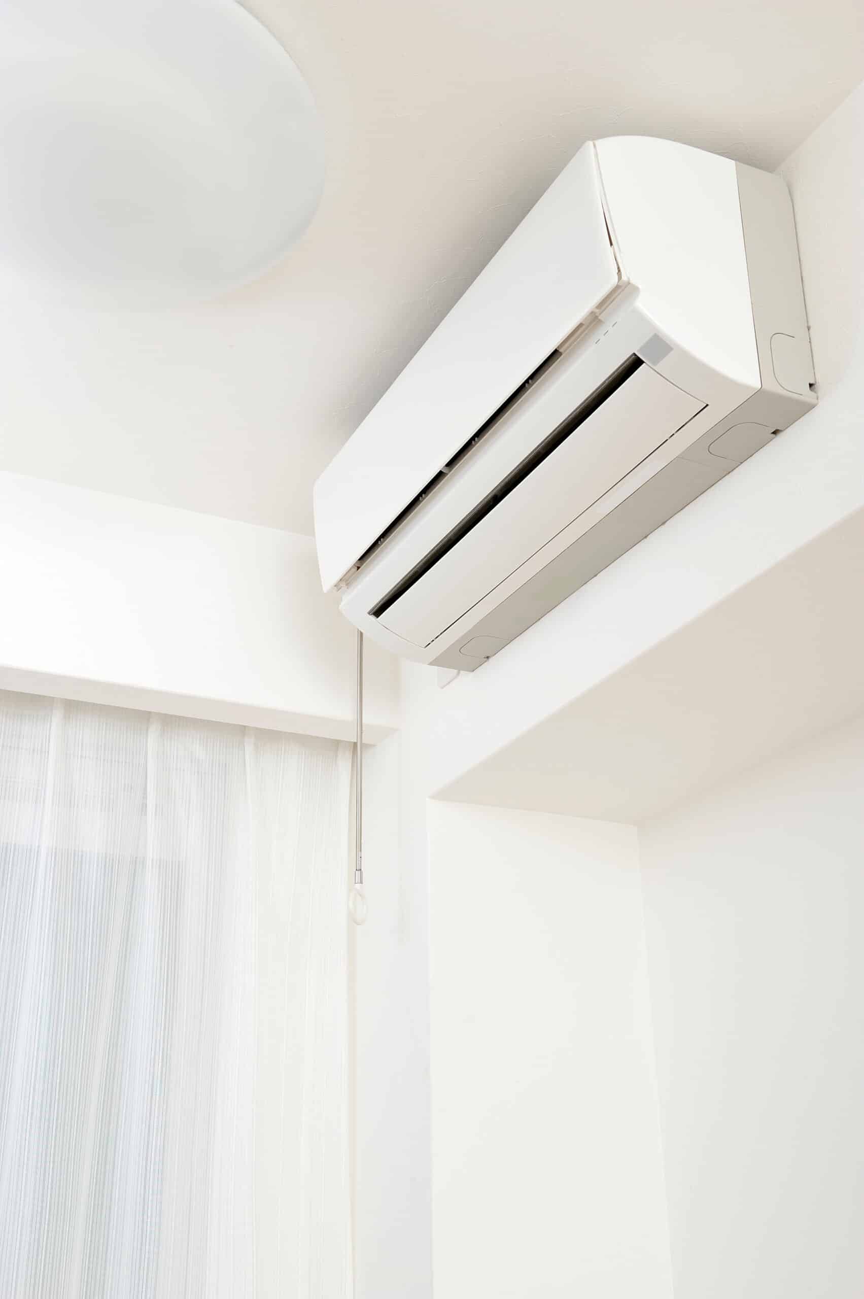 Climatiseur mural blanc installé en hauteur dans une pièce lumineuse avec plafond et murs clairs. Fenêtre avec rideaux transparents rayés.