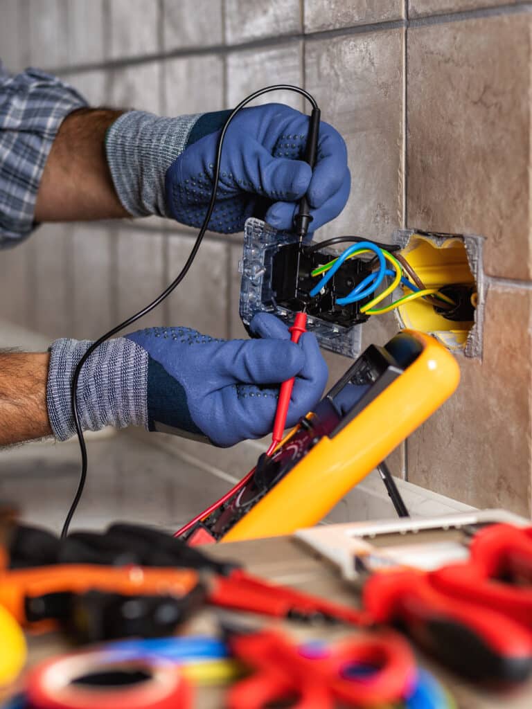 Électricien : Test et réparation de prise électrique. Électricien en gants bleus testant une prise électrique murale avec un multimètre. Des fils colorés et des outils sont visibles.
