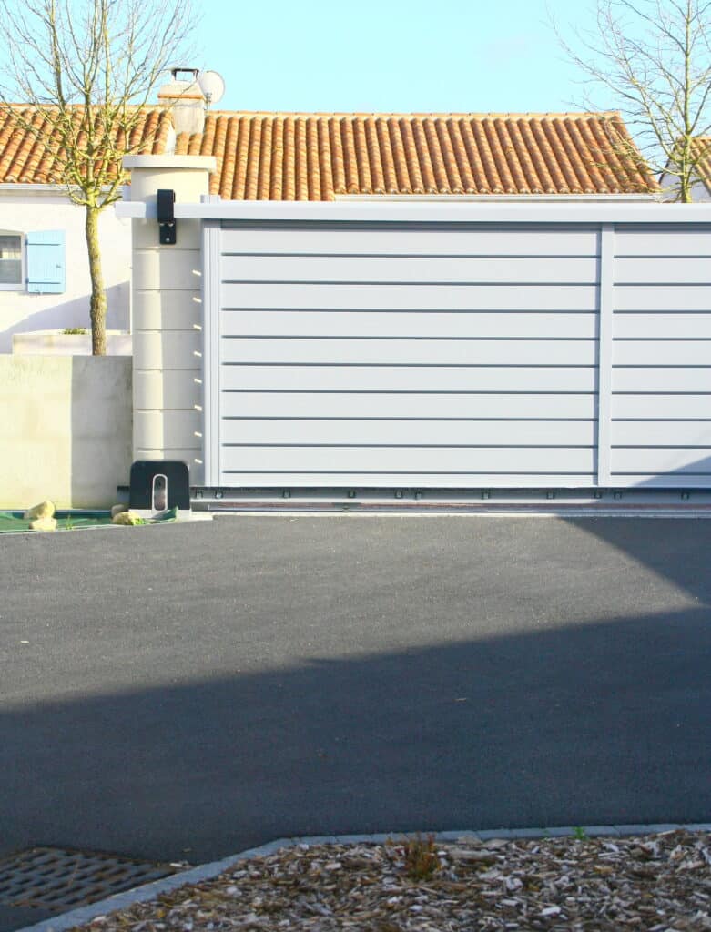 Portail coulissant automatique gris en aluminium devant une maison au toit de tuiles rouges et arbres dénudés. Allée goudronnée ombragée.