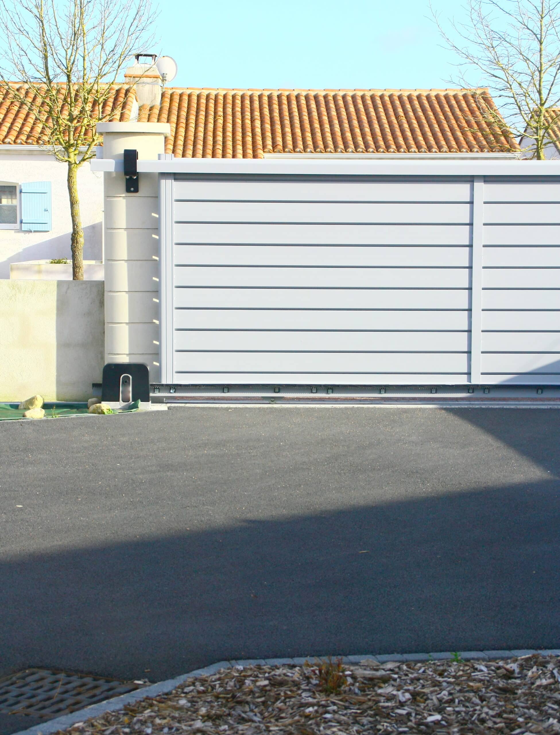 Portail coulissant automatique gris en aluminium devant une maison au toit de tuiles rouges et arbres dénudés. Allée goudronnée ombragée.